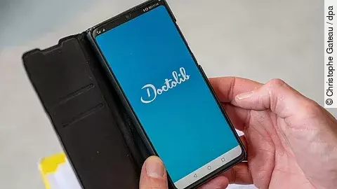 Hand hält ein Smartphone mit geöffneter Doctolib‑App, das Logo ist auf blauem Hintergrund zu sehen.