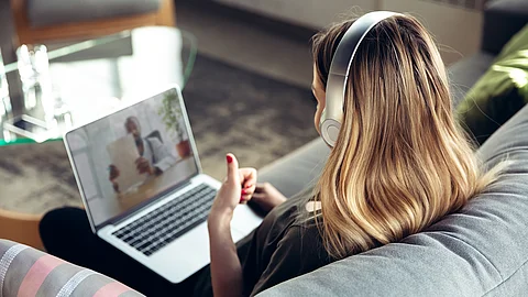 Eine Person sitzt auf einem Sofa mit einem Laptop auf dem Schoß, trägt große Kopfhörer und nimmt an einer Videositzung teil; auf dem Laptopbildschirm ist eine weitere Person zu sehen, die ein Blatt Papier hochhält.