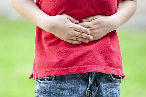 Ein Kind trägt ein rotes T-Shirt und blaue Jeans und hält sich mit beiden Händen den Bauch, was auf Bauchschmerzen hinweist.
