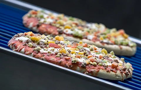 Zwei ungebackene Fertigpizzen mit Gemüse und Fleischbelag liegen auf einem blauen Förderband in einer Produktionsanlage. Zwei ungebackene Fertigpizzen mit Gemüse und Fleischbelag liegen auf einem blauen Förderband in einer Produktionsanlage.
