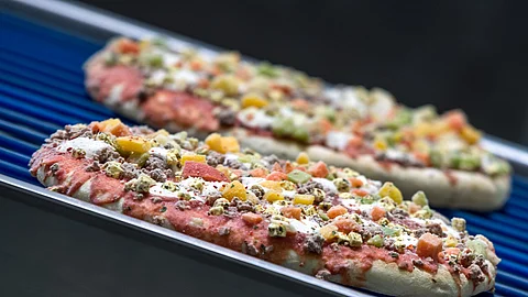 Zwei ungebackene Fertigpizzen mit Gemüse und Fleischbelag liegen auf einem blauen Förderband in einer Produktionsanlage.