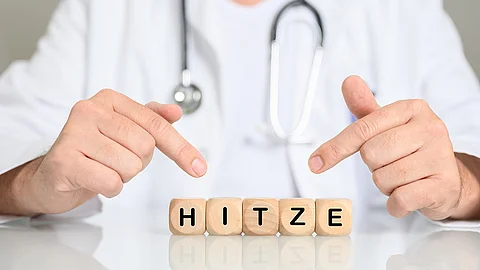 Eine Person im weißen Kittel mit Stethoskop zeigt mit beiden Zeigefingern auf Holzwürfel, auf denen das Wort „HITZE“ geschrieben steht.