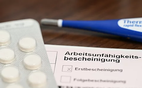 Ein Fieberthermometer, Medikamente und eine ärztliche Arbeitsunfähigkeitsbescheinigung (Krankmeldung) liegen auf einem Nachttisch 