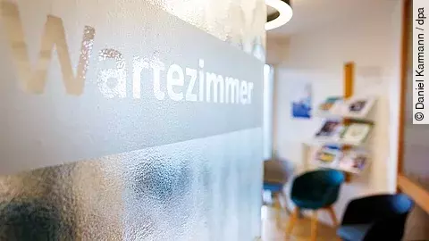 Glas-Tür mit der Aufschrift „Wartezimmer“, dahinter ein heller Raum mit Stühlen und einem Regal mit Broschüren – typisches Wartezimmer einer Arztpraxis.