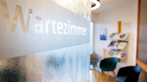 Glas-Tür mit der Aufschrift „Wartezimmer“, dahinter ein heller Raum mit Stühlen und einem Regal mit Broschüren – typisches Wartezimmer einer Arztpraxis.