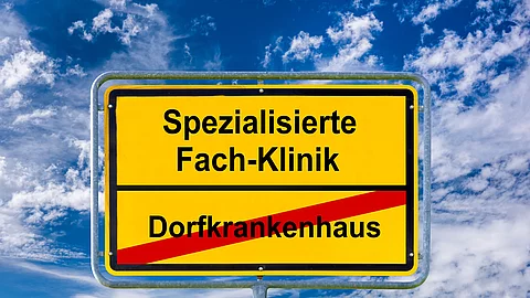 Gelbes Ortsschild vor blauem Himmel mit weißen Wolken. Auf dem Schild steht oben ‚Spezialisierte Fach-Klinik‘ in schwarzer Schrift. Darunter ist ‚Dorfkrankenhaus‘ in schwarzer Schrift rot durchgestrichen. Das Schild symbolisiert den Übergang von kleinen Krankenhäusern zu spezialisierten Kliniken.