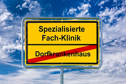 Gelbes Ortsschild vor blauem Himmel mit weißen Wolken. Auf dem Schild steht oben ‚Spezialisierte Fach-Klinik‘ in schwarzer Schrift. Darunter ist ‚Dorfkrankenhaus‘ in schwarzer Schrift rot durchgestrichen. Das Schild symbolisiert den Übergang von kleinen Krankenhäusern zu spezialisierten Kliniken.