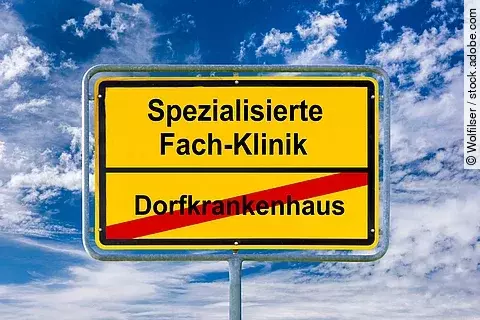Gelbes Ortsschild vor blauem Himmel mit weißen Wolken. Auf dem Schild steht oben ‚Spezialisierte Fach-Klinik‘ in schwarzer Schrift. Darunter ist ‚Dorfkrankenhaus‘ in schwarzer Schrift rot durchgestrichen. Das Schild symbolisiert den Übergang von kleinen Krankenhäusern zu spezialisierten Kliniken.