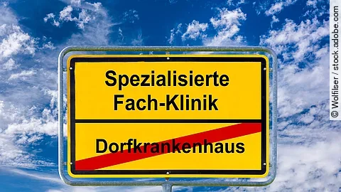 Gelbes Ortsschild vor blauem Himmel mit weißen Wolken. Auf dem Schild steht oben ‚Spezialisierte Fach-Klinik‘ in schwarzer Schrift. Darunter ist ‚Dorfkrankenhaus‘ in schwarzer Schrift rot durchgestrichen. Das Schild symbolisiert den Übergang von kleinen Krankenhäusern zu spezialisierten Kliniken.