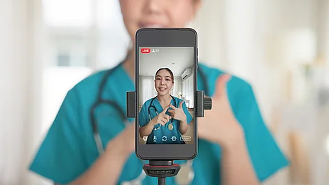in Smartphone auf einem Stativ zeigt eine Person in medizinischer Arbeitskleidung mit Stethoskop, die vor der Kamera spricht. 