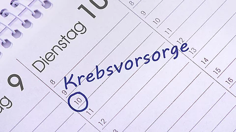 Nahaufnahme eines aufgeschlagenen Kalenders mit handschriftlichem Eintrag ‚Krebsvorsorge‘ am 10. des Monats, blau markiert. Nahaufnahme eines aufgeschlagenen Kalenders mit handschriftlichem Eintrag ‚Krebsvorsorge‘ am 10. des Monats, blau markiert.