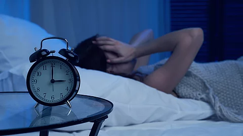 Ein analoger Wecker steht auf einem Glastisch und zeigt kurz nach 3 Uhr; im Hintergrund liegt eine Person auf einem Bett und hält den Kopf mit den Händen