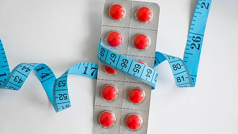 Ein Blister mit zehn roten Tabletten liegt auf hellem Hintergrund. Ein blaues Maßband ist locker darum geschlungen und zeigt Zentimeterangaben.