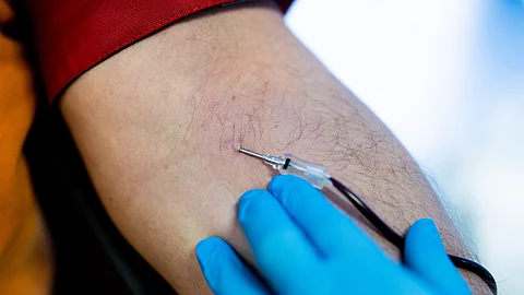 Nahaufnahme einer Blutspende: Eine Hand mit blauen Handschuhen hält eine Nadel, die in den Arm eines Spenders eingeführt ist. Ein Schlauch führt das Blut ab.
