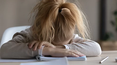 Ein Kind sitzt an einem Tisch, den Kopf auf die Arme gelegt, vor sich ein aufgeschlagenes Heft und ein Stift. Die Körperhaltung deutet auf starke Erschöpfung hin.