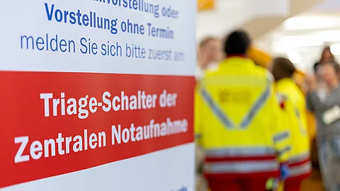 Ein Hinweisschild in einer Klinik weist auf den Triage-Schalter der zentralen Notaufnahme hin. Im Hintergrund sind zwei Personen in Rettungsdienstkleidung unscharf zu erkennen. Ein Hinweisschild in einer Klinik weist auf den Triage-Schalter der zentralen Notaufnahme hin. Im Hintergrund sind zwei Personen in Rettungsdienstkleidung unscharf zu erkennen.
