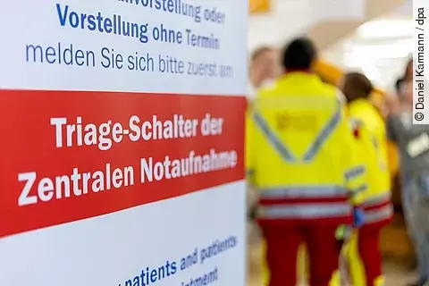 Ein Hinweisschild in einer Klinik weist auf den Triage-Schalter der zentralen Notaufnahme hin. Im Hintergrund sind zwei Personen in Rettungsdienstkleidung unscharf zu erkennen.