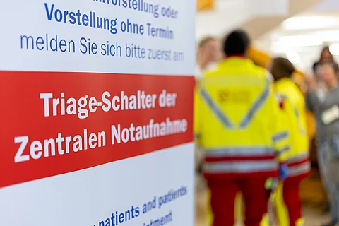 Ein Hinweisschild in einer Klinik weist auf den Triage-Schalter der zentralen Notaufnahme hin. Im Hintergrund sind zwei Personen in Rettungsdienstkleidung unscharf zu erkennen.