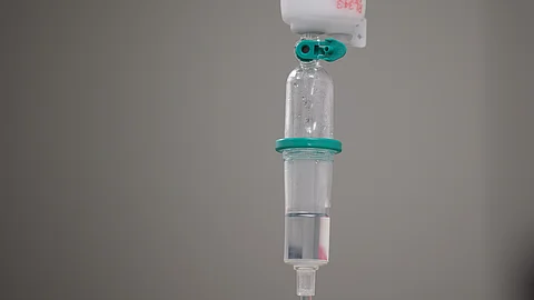 Nahaufnahme eines medizinischen Infusionssystems mit transparentem Behälter, Schlauch und Anschluss, aus dem Flüssigkeit nach unten läuft.