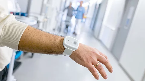 Nahaufnahme eines Armes mit weißem Patientenarmband, das einen Bluetooth-Transponder enthält. Im Hintergrund ein Krankenhausflur mit unscharfen Personen.