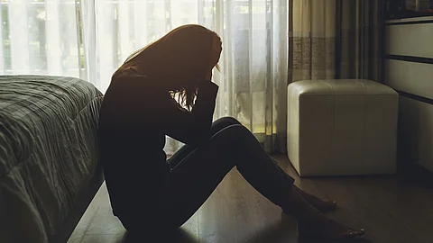 Eine junge Frau sitzt mit dem Kopf in den Händen in einem dunklen Schlafzimmer auf dem Fußboden