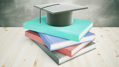 Ein Stapel mehrfarbiger Bücher liegt auf einem Holztisch, darauf befindet sich ein akademischer Doktorhut vor einem unscharfen Tafelhintergrund.