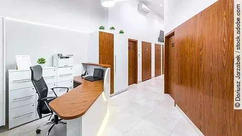 Ein heller, sauber strukturierter Empfangsbereich einer Arztpraxis mit mehreren Türen zu Behandlungszimmern, einem Schreibtisch mit Monitor und einem übersichtlichen, modernen Interieur.